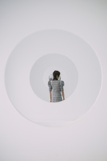 A person stands facing away, positioned centrally within a series of concentric circular frames giving a surreal, tunnel-like visual effect. The individual is wearing a plaid dress, contributing a vintage or classic tone to the minimalist and futuristic setting.