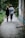 A young couple is walking hand in hand down a narrow, tree-lined path beside a brick building. The ground is paved and surrounded by lush greenery, suggesting a peaceful outdoor setting.