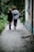 A young couple is walking hand in hand down a narrow, tree-lined path beside a brick building. The ground is paved and surrounded by lush greenery, suggesting a peaceful outdoor setting.