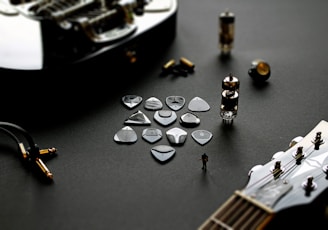 A collection of guitar picks is laid out on a dark surface, accompanied by various guitar components and accessories, including guitar strings, a tube amplifier, and a guitar headstock. The setup conveys a sense of musical creation and technical assembly.
