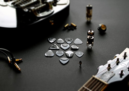 A collection of guitar picks is laid out on a dark surface, accompanied by various guitar components and accessories, including guitar strings, a tube amplifier, and a guitar headstock. The setup conveys a sense of musical creation and technical assembly.