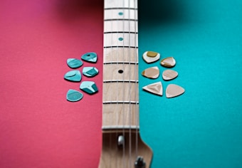A guitar neck is placed diagonally across a two-toned background split into red on the left and teal on the right. On each side of the neck, clusters of guitar picks are arranged. The picks on the red side are teal, while the picks on the teal side are beige, creating a contrasting color scheme.