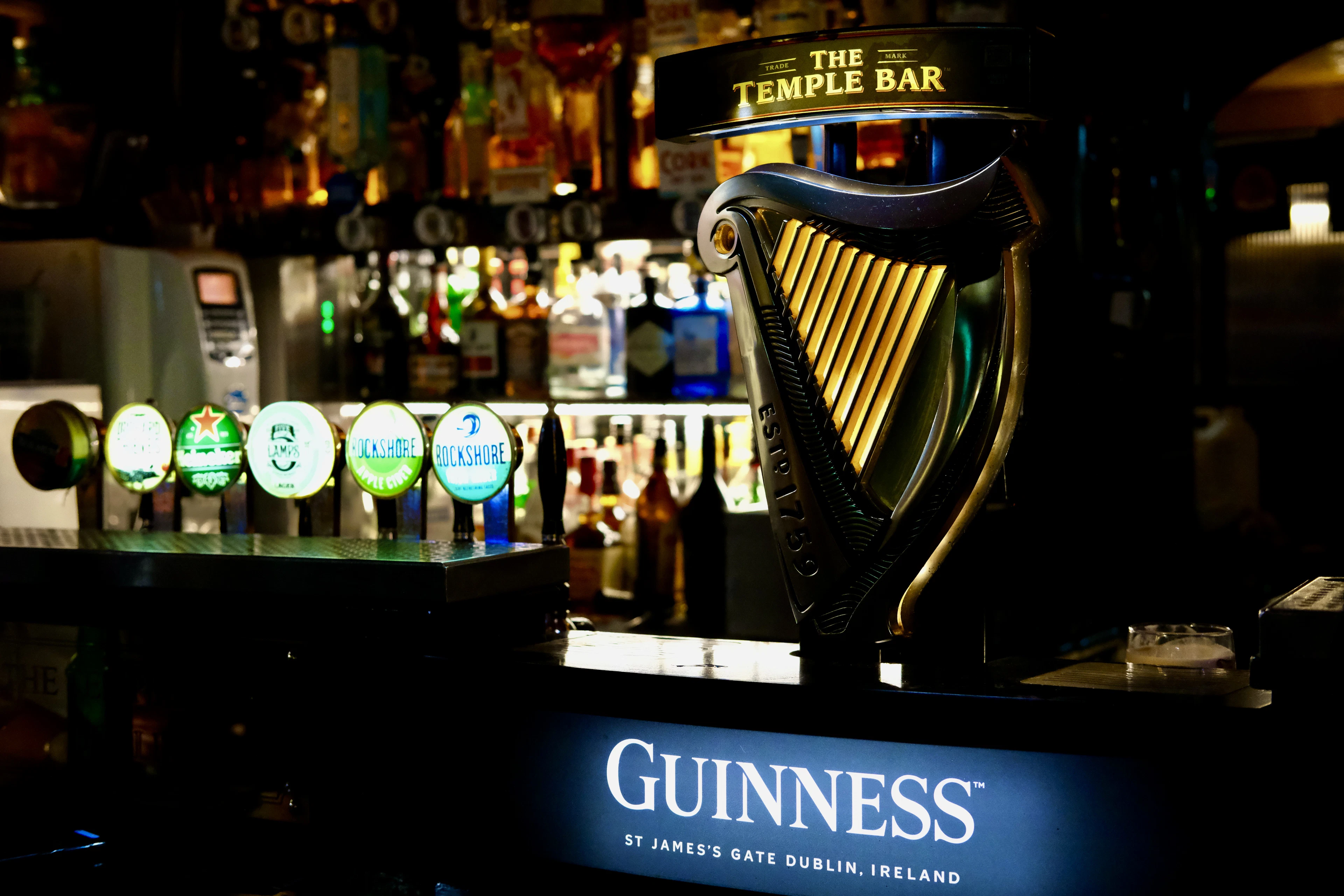 A brass Guinness harp tap on the bar of The Temple Bar pub in Dublin, backlit bottles and taps behind it