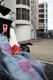 A street scene with tall modern apartment buildings in the background and several garbage bags and a trash container in the foreground. The trash bags are gray with pink designs on them, and there is an overturned red crate among the refuse. The buildings are multi-storied with balconies and large windows, and the area appears to be an urban setting.