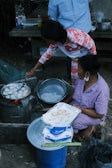 Two people are engaged in cooking street food outdoors. One person skillfully manages a pan on a makeshift stove, preparing small, round dough pieces. The other person, seated, holds a tray lined with paper and freshly cooked items. Various kitchen supplies and condiments, including bottles and bowls, are around them. The setting suggests an informal, communal cooking space.