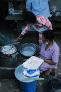 Two people are engaged in cooking street food outdoors. One person skillfully manages a pan on a makeshift stove, preparing small, round dough pieces. The other person, seated, holds a tray lined with paper and freshly cooked items. Various kitchen supplies and condiments, including bottles and bowls, are around them. The setting suggests an informal, communal cooking space.
