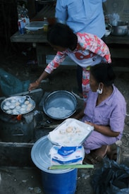 Two people are engaged in cooking street food outdoors. One person skillfully manages a pan on a makeshift stove, preparing small, round dough pieces. The other person, seated, holds a tray lined with paper and freshly cooked items. Various kitchen supplies and condiments, including bottles and bowls, are around them. The setting suggests an informal, communal cooking space.
