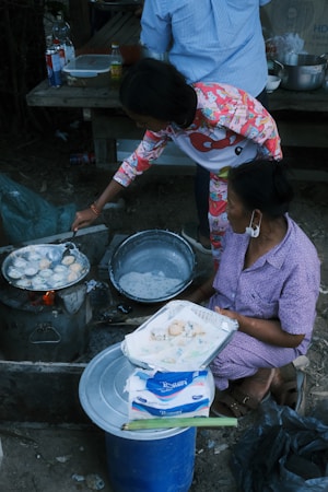 Two people are engaged in cooking street food outdoors. One person skillfully manages a pan on a makeshift stove, preparing small, round dough pieces. The other person, seated, holds a tray lined with paper and freshly cooked items. Various kitchen supplies and condiments, including bottles and bowls, are around them. The setting suggests an informal, communal cooking space.