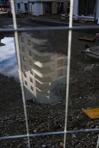 A reflection of a building with balconies and windows is visible in a puddle on a construction site. The foreground is filled with gravel and some construction materials, while a fence partially obscures the view.