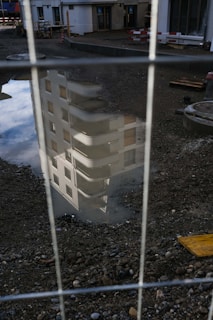 A reflection of a building with balconies and windows is visible in a puddle on a construction site. The foreground is filled with gravel and some construction materials, while a fence partially obscures the view.