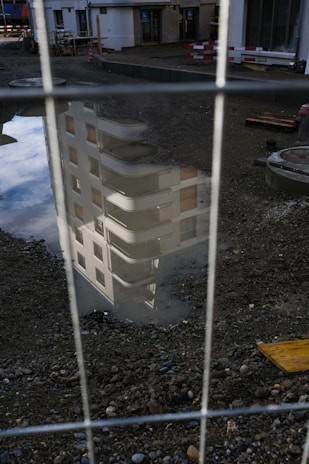 A reflection of a building with balconies and windows is visible in a puddle on a construction site. The foreground is filled with gravel and some construction materials, while a fence partially obscures the view.