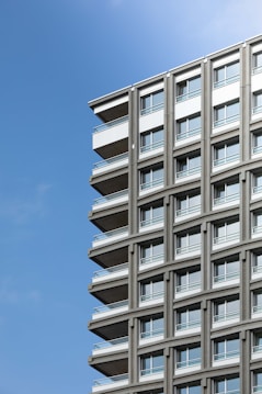 A modern high-rise building with a repeating pattern of windows and balconies, positioned against a clear blue sky. The architecture features clean lines and neutral colors, enhancing its contemporary design aesthetic.