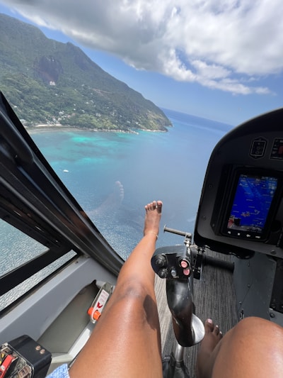 A person with legs extended rests inside a helicopter, overlooking a scenic coastal landscape. The view captures a turquoise sea and a lush green mountain under a partly cloudy sky. The helicopter cockpit interior is partially visible, including control sticks and a display screen.