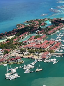An aerial view of a coastal residential area with numerous red-roofed buildings interspersed with lush green trees. A marina is populated with various boats and yachts floating in the turquoise water. The surrounding sea is a clear blue, transitioning from light to dark shades, and a small section of coral reef is visible.