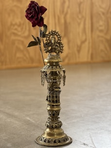 A brass decorative object featuring intricate designs and motifs, standing upright on a smooth surface. At the top of the object, there is a detailed sculpture of a deity, likely from Hindu iconography, with a halo behind it. A single deep red rose is positioned to appear as if growing from the top of the sculpture, adding contrast. The background is a wooden, textured panel.