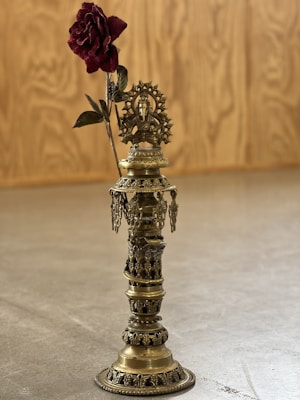 A brass decorative object featuring intricate designs and motifs, standing upright on a smooth surface. At the top of the object, there is a detailed sculpture of a deity, likely from Hindu iconography, with a halo behind it. A single deep red rose is positioned to appear as if growing from the top of the sculpture, adding contrast. The background is a wooden, textured panel.
