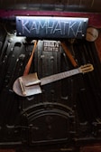 An industrial-looking black metal surface with a handmade sign reading 'Камчатка' in blue above it. A metallic shovel is creatively modified into a guitar, hanging from leather straps across the surface. A small metal gauge and additional markings are visible.