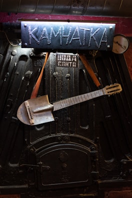 An industrial-looking black metal surface with a handmade sign reading 'Камчатка' in blue above it. A metallic shovel is creatively modified into a guitar, hanging from leather straps across the surface. A small metal gauge and additional markings are visible.