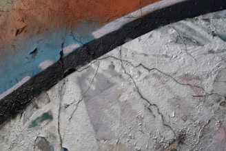 A close-up view of a textured wall with cracked paint. The wall features abstract graffiti with strokes of blue, white, and orange, layered over a white and black background. The surface shows signs of aging, with visible cracks and peeled areas.
