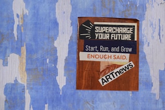 A weathered blue wall with peeling paint has a poster attached. The poster features text with motivational phrases such as 'Supercharge Your Future', 'Start, Run, and Grow', and 'Enough Said'. It includes the logo for ARTnews.
