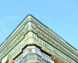 An upward view of a green building with multiple floors and large windows against a clear blue sky. The architectural style features prominent green wooden structures surrounding the windows.