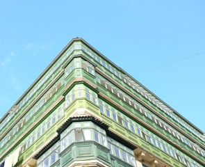 An upward view of a green building with multiple floors and large windows against a clear blue sky. The architectural style features prominent green wooden structures surrounding the windows.