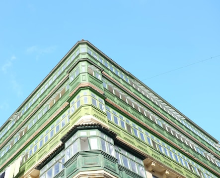 An upward view of a green building with multiple floors and large windows against a clear blue sky. The architectural style features prominent green wooden structures surrounding the windows.