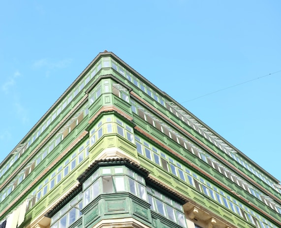 An upward view of a green building with multiple floors and large windows against a clear blue sky. The architectural style features prominent green wooden structures surrounding the windows.