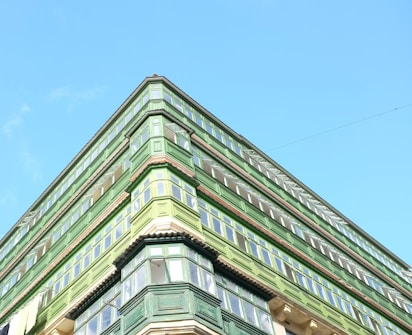 An upward view of a green building with multiple floors and large windows against a clear blue sky. The architectural style features prominent green wooden structures surrounding the windows.
