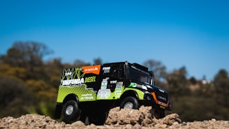 A toy truck is positioned on a mound of dirt with detailed branding and logos. The truck seems designed to resemble an off-road or rally vehicle, with vibrant colors and sponsor decals. The background features a soft-focus landscape with trees under a clear blue sky.