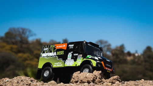 A toy truck is positioned on a mound of dirt with detailed branding and logos. The truck seems designed to resemble an off-road or rally vehicle, with vibrant colors and sponsor decals. The background features a soft-focus landscape with trees under a clear blue sky.