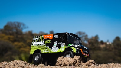 A toy truck is positioned on a mound of dirt with detailed branding and logos. The truck seems designed to resemble an off-road or rally vehicle, with vibrant colors and sponsor decals. The background features a soft-focus landscape with trees under a clear blue sky.