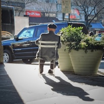An elderly person wearing a cap sits on a white chair on an urban sidewalk. The individual is facing away, with a black SUV parked in front and a large potted plant beside them. The shadow of the person and chair is cast on the ground, and signs from the UPS Store and RadioShack are visible in the background.