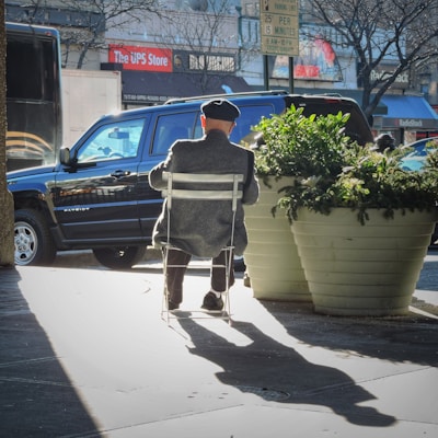 An elderly person wearing a cap sits on a white chair on an urban sidewalk. The individual is facing away, with a black SUV parked in front and a large potted plant beside them. The shadow of the person and chair is cast on the ground, and signs from the UPS Store and RadioShack are visible in the background.