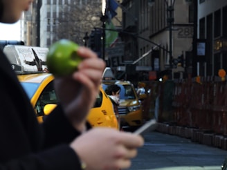 A city street scene featuring a person holding a green apple and a smartphone in the foreground. Yellow taxis are visible in the background, along with a busy urban environment with buildings and advertisements. The focus is primarily on the apple and the urban setting.