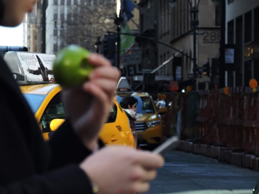 A city street scene featuring a person holding a green apple and a smartphone in the foreground. Yellow taxis are visible in the background, along with a busy urban environment with buildings and advertisements. The focus is primarily on the apple and the urban setting.