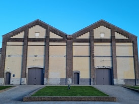 A large, symmetrical brick building with an industrial look features two peaks and a series of rectangular patterns. The bricks vary in color from light beige to dark brown, creating a contrast. Large wooden doors are centrally located on both sections of the building, with smaller entranceways on each side. The foreground shows a manicured lawn with a small path leading up to the building. The sky is clear and vibrant blue.