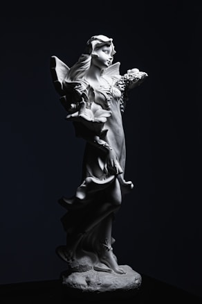 A detailed statue of a graceful female figure depicting an angelic or fairy-like character. The statue features intricate details such as flowing hair, wings, and a long flowing gown. The lighting highlights the fine craftsmanship, casting shadows that enhance the texture and form.