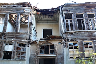 A dilapidated, abandoned house with a severely damaged roof and broken windows. The wooden structure shows signs of decay, with missing planks and exposed beams. Vegetation is growing at the base, adding to the sense of neglect and abandonment.