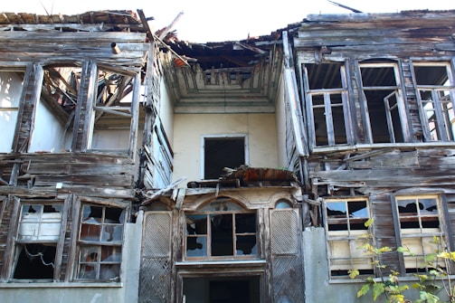 A dilapidated, abandoned house with a severely damaged roof and broken windows. The wooden structure shows signs of decay, with missing planks and exposed beams. Vegetation is growing at the base, adding to the sense of neglect and abandonment.
