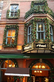 A storefront decorated with festive garlands and lights. The building features brick architecture and large windows. Greenery and red ornaments adorn the facade, while warm light emanates from inside, creating a cozy atmosphere. A canopy displays the name 'VITAVIEN' in white letters.