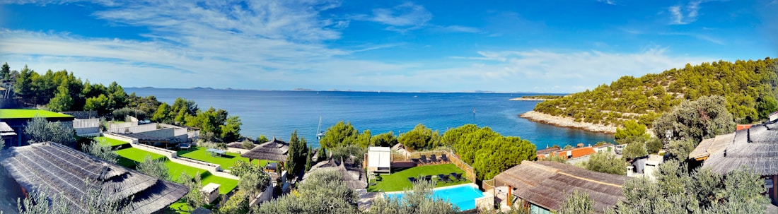 A serene coastal landscape with clear blue skies and calm blue sea. The foreground features resort-like accommodations with green lawns, a swimming pool, and wooden structures. Lush green trees and vegetation are scattered throughout the area, creating a contrast with the blue water. A rocky, tree-covered hill extends into the sea to the right.