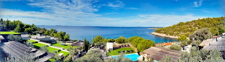 A serene coastal landscape with clear blue skies and calm blue sea. The foreground features resort-like accommodations with green lawns, a swimming pool, and wooden structures. Lush green trees and vegetation are scattered throughout the area, creating a contrast with the blue water. A rocky, tree-covered hill extends into the sea to the right.
