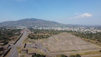 Aerial view of an ancient archaeological site with large pyramidal structures amid a vast landscape. The ruins are surrounded by a flat plain that extends towards a mountain range in the background. Scattered trees and small structures fill the area, while a modern cityscape is visible at the base of the mountains.