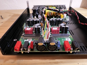 A detailed view of an electronic circuit board with various components including capacitors, resistors, connectors, and colored wiring. The board is housed within a metallic or plastic case, and includes multiple red and black terminal connectors, as well as larger circular connectors.