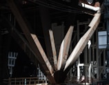 A close-up view of large, rusted steel beams forming part of a structural support, with dark shadows and patches of light. The beams are intersecting and have engraved markings. Background structures are visible, suggesting an industrial or construction setting.