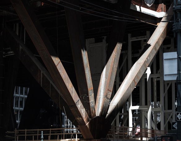 A close-up view of large, rusted steel beams forming part of a structural support, with dark shadows and patches of light. The beams are intersecting and have engraved markings. Background structures are visible, suggesting an industrial or construction setting.