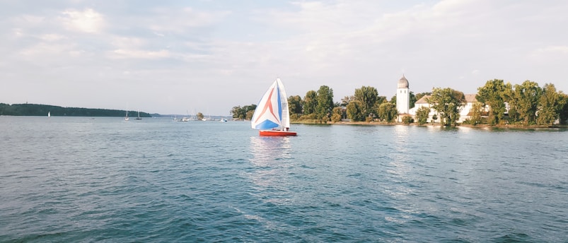 A small sailboat with a colorful red, white, and blue sail is gliding on a calm body of water. In the background, there is a scenic landscape featuring a cluster of trees and a church with a domed tower along the shoreline. The sky is partly cloudy, casting a serene atmosphere over the scene.