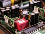 A close-up view of electronic components on a circuit board, featuring capacitors, resistors, and microchips. The arrangement includes cylindrical capacitors alongside rectangular chips with QR codes and other components connected by intricate wiring.