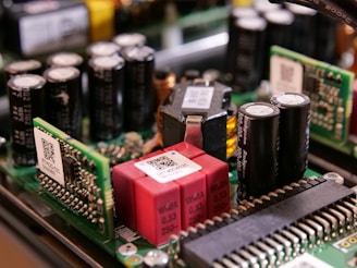 A close-up view of electronic components on a circuit board, featuring capacitors, resistors, and microchips. The arrangement includes cylindrical capacitors alongside rectangular chips with QR codes and other components connected by intricate wiring.